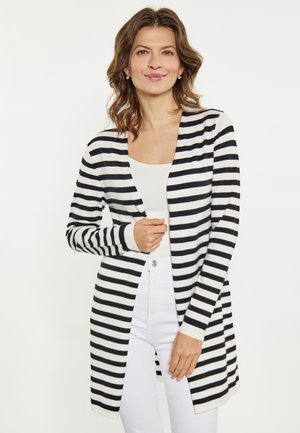 Frau trägt einen schwarz-weiß horizontal gestreiften Cardigan über weißem Top und weißen Hosen, lächelt vor hellem einfarbigem Hintergrund.
