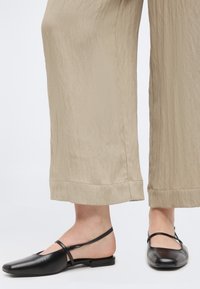 Pieds portant des ballerines en cuir noir à bride arrière avec des lanières croisées, associées à un pantalon large beige et ample.
