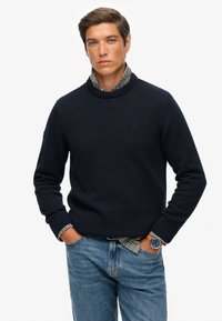 Homme portant un pull marine sur une chemise à carreaux, avec un jean bleu et une montre, se tenant les mains dans les poches contre un fond blanc.