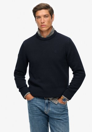 Man in een marineblauwe trui over een geblokt shirt, met blauwe spijkerbroek en een horloge, staande met zijn handen in zijn zakken tegen een witte achtergrond.