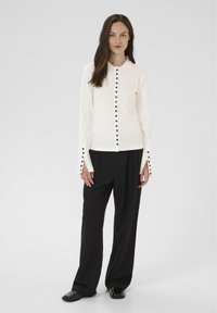 Kobieta stojąca w białej bluzce z guzikami na długim rękawie i czarnymi guzikami oraz w luźnych czarnych spodniach na tle białego tła.