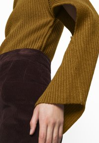 Sininen ribatut neulepaidat leveillä hihoilla, yhdistetty tummanruskeaan nyörikankaiseen hameeseen, jossa on pystysuorat raidat. Lähikuva kankaan tekstuureista.