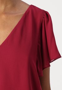 Burgundy blouse with a v-neckline and short, draped sleeves. Made from lightweight fabric with a smooth texture. Accented shoulder gathering.
