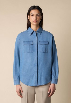 Kobieta o prostych brązowych włosach, ubrana w niebieską jeansową koszulę z dwoma kieszeniami na piersi oraz beżowe spodnie, stojąca na jednolitym tle.