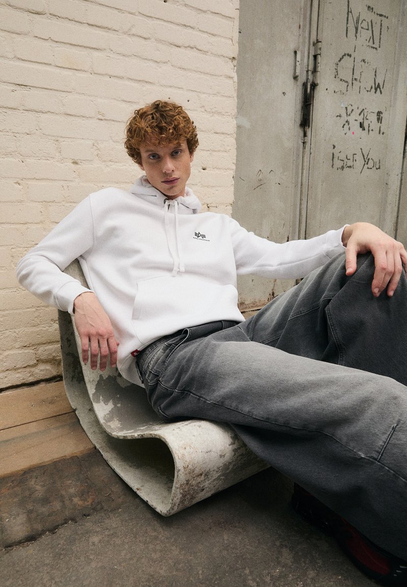 Witte hoodie met een voorkantzak en logo, gecombineerd met grijze baggy jeans. Model zit op een gebogen betonsculptuur tegen een gestructureerde muur.
