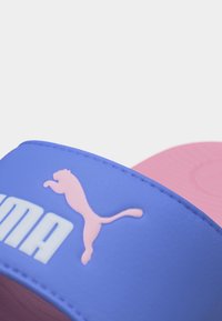 Κοντινό πλάνο του λουριού της παντόφλας Puma σε μωβ και ροζ χρώμα με ροζ λογότυπο Puma και λευκά γράμματα σε απλό φόντο.