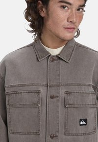 Graue Jeansjacke mit Kragen, ausgestattet mit zwei Fronttaschen, braunen Knopfverschlüssen und einem kleinen Logo-Patch auf der linken Tasche.