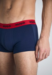 Námornícky modré boxerky s červeným elastickým pásom, na ktorom je v čiernej farbe nápis "HUGO". Tkanina vyzerá hladko a má priliehavý strih.