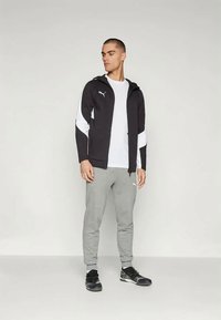 Czarna kurtka zapinana na zamek z białymi akcentami, w połączeniu z szarymi dresami. Strój charakteryzuje się sportowym stylem i logo Puma na każdej z części.