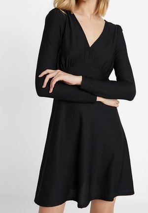 Femme portant une robe noire à manches longues avec un décolleté en V et une taille cintrée, debout les bras croisés devant un fond uni.