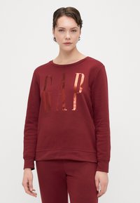 Bordeaux bluzа z długim rękawem wykonana z mieszanki bawełny, z metalicznym logo "GAP" w nowoczesnej czcionce. Okrągły dekolt. Gładka tekstura.