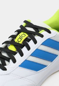 Scarpa sportiva bianca con strisce blu e lacci neri. Presenta una texture a rete, collo imbottito e un'etichetta logo gialla brillante.