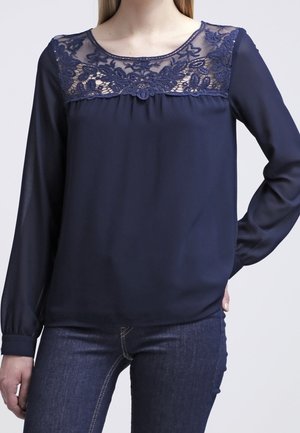 Frau trägt eine marineblaue langärmelige Bluse mit floraler Spitzenverzierung am Ausschnitt und dunkelblaue Jeans, Hand in die Hüfte gestützt.