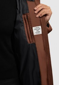 Brūnā quiltēta žakete ar melnu oderi, sānu kabatu un etiķeti ar tekstu "Silts apskāviens, kas izgatavots no auduma."