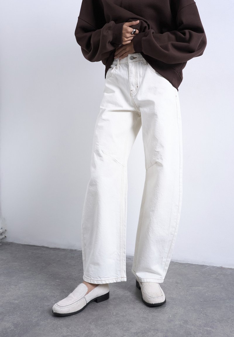 Άτομο που φοράει λευκό παντελόνι με φαρδιά μπατζάκια, λευκά παπούτσια χωρίς κορδόνια και ένα υπερμεγέθες σκούρο καφετί πουλόβερ, στέκεται σε γκρι πάτωμα μπροστά σε λευκό τοίχο.