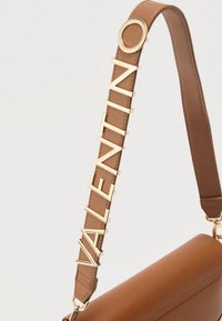 Brązowy pasek ze skóry do torby, z złotymi literami tworzącymi napis "VALENTINO", przymocowany do beżowej torby z ziarnistej skóry.