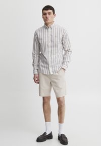 Knapplang skjorte med tynne multikolor stripe, krage og lange ermer; kombinert med beige shorts og mørke slip-on sko. Hvite sokker synlige.