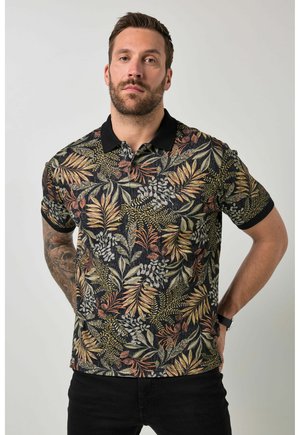 Man met baard die een zwart poloshirt draagt met een patroon van herfstbladeren, staand met de handen achter zijn rug tegen een effen achtergrond.