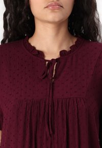 Femme portant un chemisier bordeaux texturé avec un col volanté et un nœud noué au centre devant, visible du cou jusqu'au milieu de la poitrine.