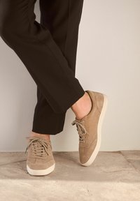 Beige semišùvne tenisky s nízkým profilem, kulatou špičkou a odpovídajícími tkaničkami. Podešev je bílá gumová, což kontrastuje s svrchním materiálem.