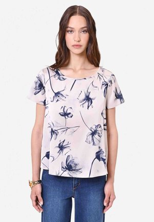 Donna che indossa una camicetta rosa chiaro a maniche corte con stampa floreale blu e jeans blu, in piedi davanti a uno sfondo bianco semplice.