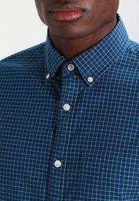 Chemise à carreaux bleue et verte avec un col boutonné, comprenant des boutons blancs, une poche poitrine et une texture lisse.