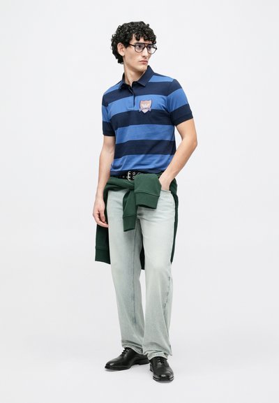 Μπλε και ναυτικό ριγέ πόλο πουκάμισο με πατς, ανοιχτόχρωμο τζιν, πράσινο φούτερ δεμένο στη μέση και μαύρα παπούτσια.