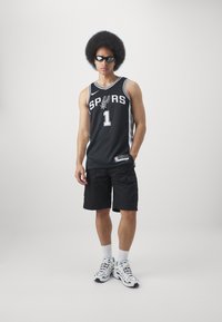 Juoda krepšinio marškinėliai su užrašu "SPURS" ir numeriu "1", derinami su juodomis kargo šortais ir baltais sportbačiais. Modelis dėvi akinius nuo saulės.