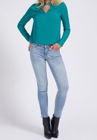 Blusa a maniche lunghe color teal con scollatura a V, abbinata a jeans skinny blu chiaro e stivali neri alla caviglia con punta affusolata.