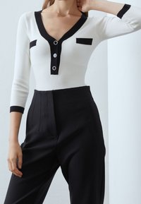 Maglia bianca a maniche lunghe con coste, scollatura a V nera, due tasche quadrate nere e dettagli con bottoni argentati; abbinata a pantaloni neri a vita alta.