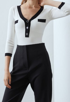 Haut à manches longues blanc à côtes avec un col en V noir, deux poches carrées noires et des accents de boutons en argent ; associé à un pantalon noir taille haute.