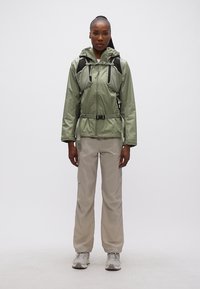 Γυναίκα που φοράει πράσινο μπουφάν με κουκούλα και ενσωματωμένο σακίδιο πλάτης, μπεζ παντελόνι cargo και γκρι αθλητικά παπούτσια, στέκεται μπροστά σε λευκό φόντο.