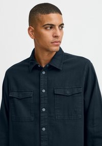Dark denim shirt featuring a button-up front, two chest pockets, and a fold-down collar. Notable gray buttons and subtle logo detail.