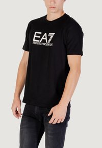 Camiseta de algodón negra con mangas cortas, que presenta un logo blanco "EA7 EMPORIO ARMANI" en el pecho. Diseño clásico de cuello redondo.