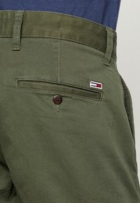 Oliivinvihreät housut, joissa on takatasku, ruskealla napilla kiinnitys ja pieni brodeerattu logo punaisena, valkoisena ja sinisenä.