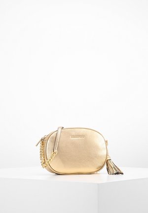 Petit sac bandoulière en cuir doré avec une bandoulière chaîne et un détail de gland, présenté sur un piédestal blanc sur un fond uni.