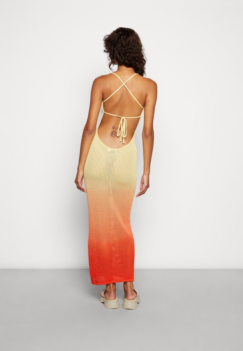 Femme debout de dos, portant une robe en maille sans manches à dos ouvert dégradée du jaune au rouge, avec des sandales compensées beige.