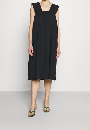Robe noire sans manches avec décolleté carré, détails froncés aux épaules et silhouette midi, fabriquée en tissu léger. Tongs vertes.