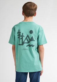 Tricou verde din bumbac, cu un design grafic pe spate, reprezentând copaci, munți și un soare, iar numele brandului scris cu un font mai mic.