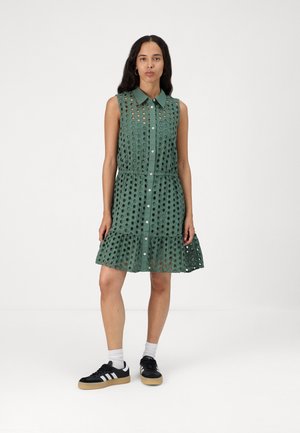 Vestido verde sin mangas con patrón de ojales, parte delantera abotonada y dobladillo con volantes; combinado con zapatillas negras y calcetines blancos hasta el tobillo.