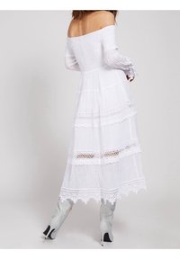 Robe maxi blanche à épaules dénudées avec corsage smocké, manches longues et bordure en dentelle superposée. Associée à des bottes argentées jusqu'aux genoux.
