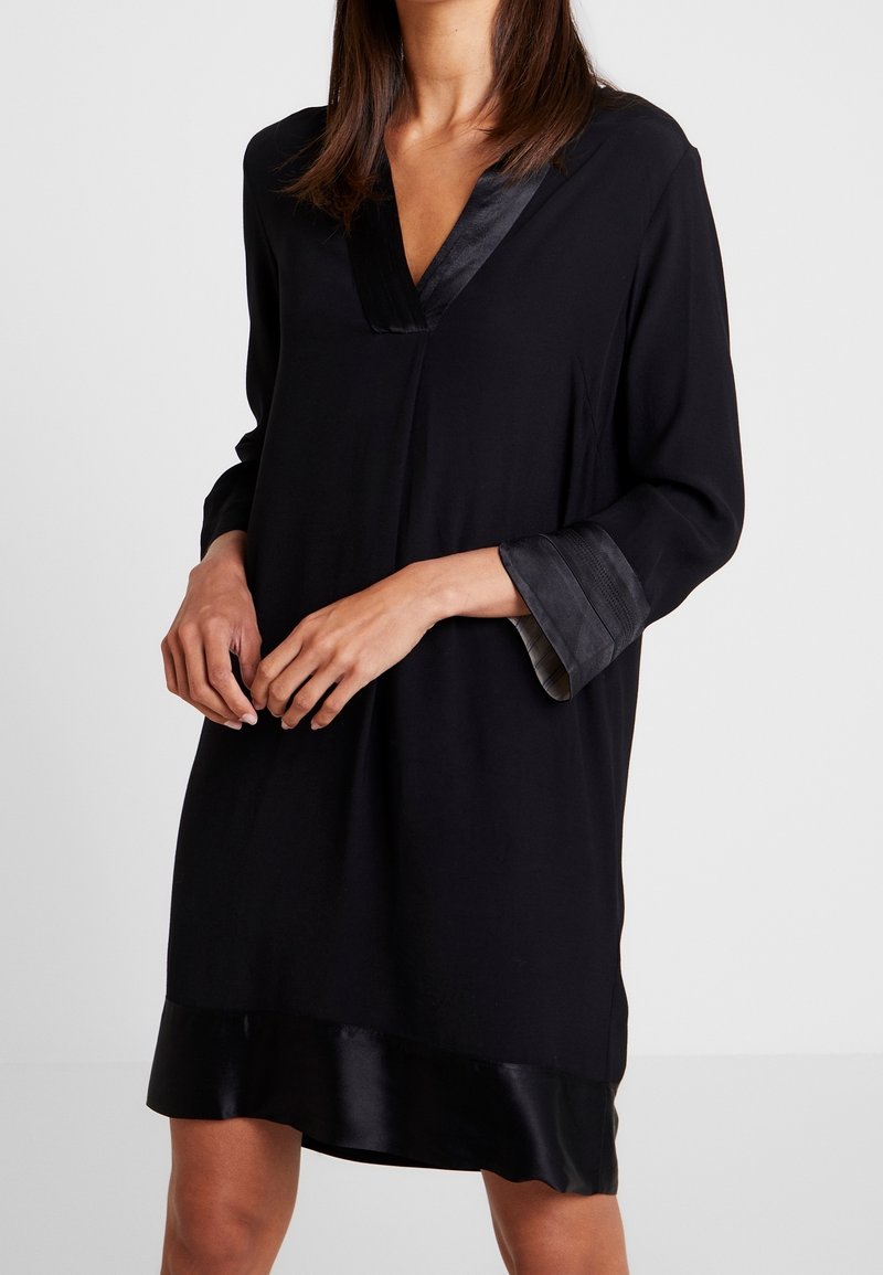 Femme portant une robe noire à manches longues avec des bordures en satin au col en V, aux poignets et à l'ourlet, se tenant devant un fond blanc uni.