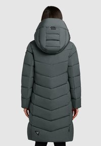 Een persoon in een lange, donkergrijze gewatteerde winterjas met een grote capuchon, gezien vanuit de rug tegen een effen achtergrond.
