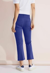 Blå utsvängda byxor i stretchigt material, med två bakfickor. Kombinerat med vita sneakers, bakgrund i mjuka pastellfärger.