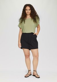 Olivenfarbene Bluse mit lockerer Passform, kombiniert mit schwarzen Shorts mit Seitentaschen und einer glatten Textur. Schwarze Sandalen mit Riemen runden das Outfit ab.