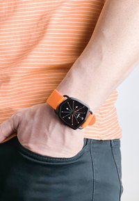 Digitalur med en sort hexagonal urskive, hvide mærker og et orange silikonearmbånd. Båret på et håndled, mod en stribet orange skjorte.