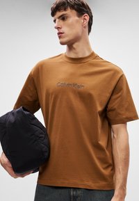 Καφέ βαμβακερό μπλουζάκι με κεντητό λογότυπο "Calvin Klein", κοντά μανίκια και χαλαρή εφαρμογή. Το μοντέλο κρατάει μια σκούρα πουφερ τσάντα.