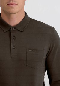 Dunkelgrünes Poloshirt aus strukturiertem Stoff, mit drei Knöpfen und einer Brusttasche, die mit einer Logo-Stickerei in kontrastierender Farbe versehen ist.