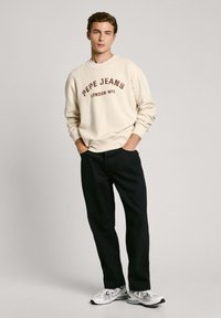 Sudadera crema con estampado "PEPE JEANS LONDON W11", combinada con pantalones oscuros de color marino y zapatillas deportivas blancas. Tejido suave, corte casual.