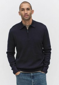 Donkerblauwe gebreide pullover met een ritskraag, geribde manchetten en zoom. Gedragen over een lichtgekleurde shirt, gecombineerd met blauwe spijkerbroek.